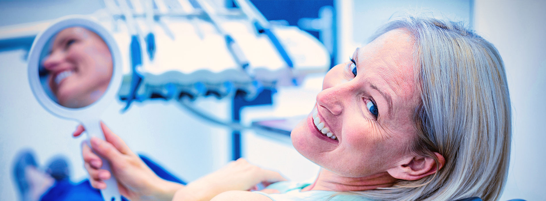 A woman is sitting in a dental chair, smiling at the camera while holding a mirror.