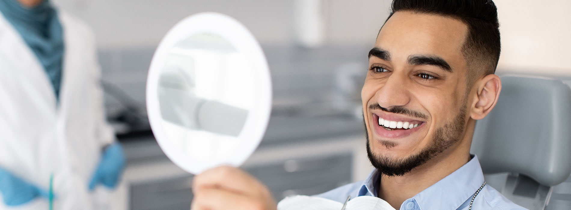 The image shows a man with a beard, smiling and holding a magnifying glass close to his face. He is wearing a blue shirt and appears to be in a dental or medical setting, as suggested by the presence of medical equipment and a person in a white coat in the background.