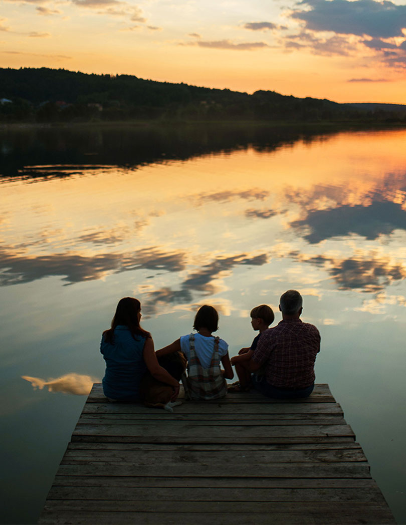 A family of four is sitting on a wooden dock at sunset, with the calm water reflecting the warm hues of the sky.