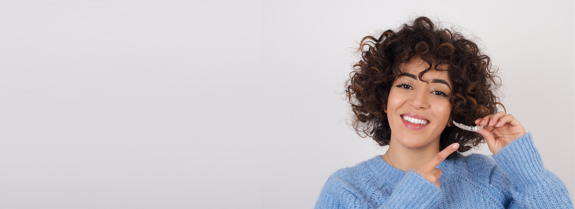 The image depicts a person with curly hair, smiling and holding a smartphone. They are wearing a blue sweater and standing against a plain background.