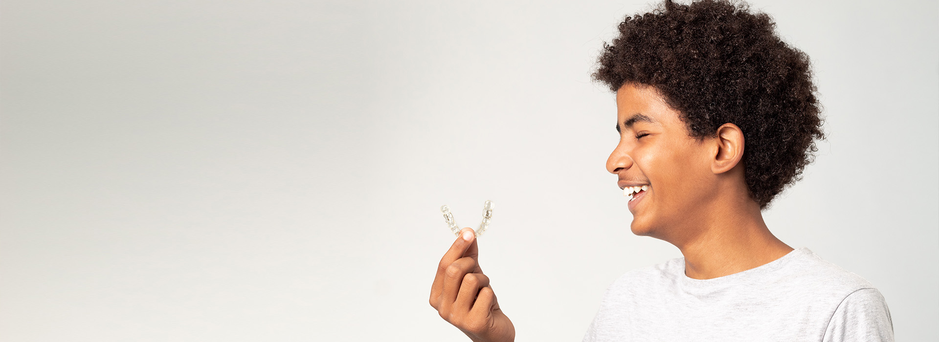 The image shows a young person smiling and holding a small white object, possibly a flower or a piece of paper, against a plain background.