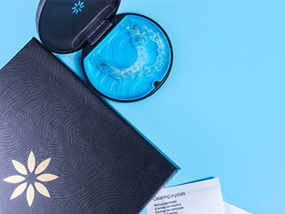 The image shows a blue box with a clear lid and a toothbrush inside, placed on a surface next to an open book or brochure with a patterned cover.