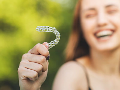 A woman is holding a clear plastic smiley with a tooth-shaped hole in the center, against a blurred outdoor background.
