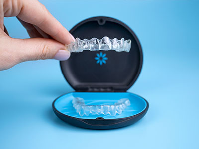 An image of a person's hand holding an open clear case containing a set of transparent dental aligners, with a blue background.
