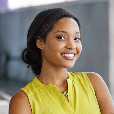 A young woman with a bright smile, wearing a yellow top and posing against a blurred background.