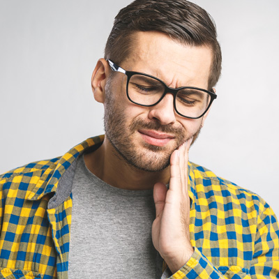 A man with glasses and a beard, wearing a plaid shirt, is captured in a moment of concern or pain with his hand on his chin.