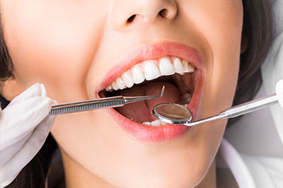 A woman with a wide smile, showcasing her teeth, in front of a dental clinic setting with a dentist's chair and tools.