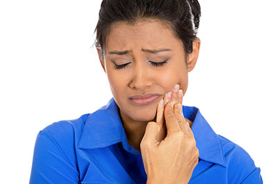 A woman in a blue shirt, holding her face with one hand and looking downward with an expression of concern or pain.