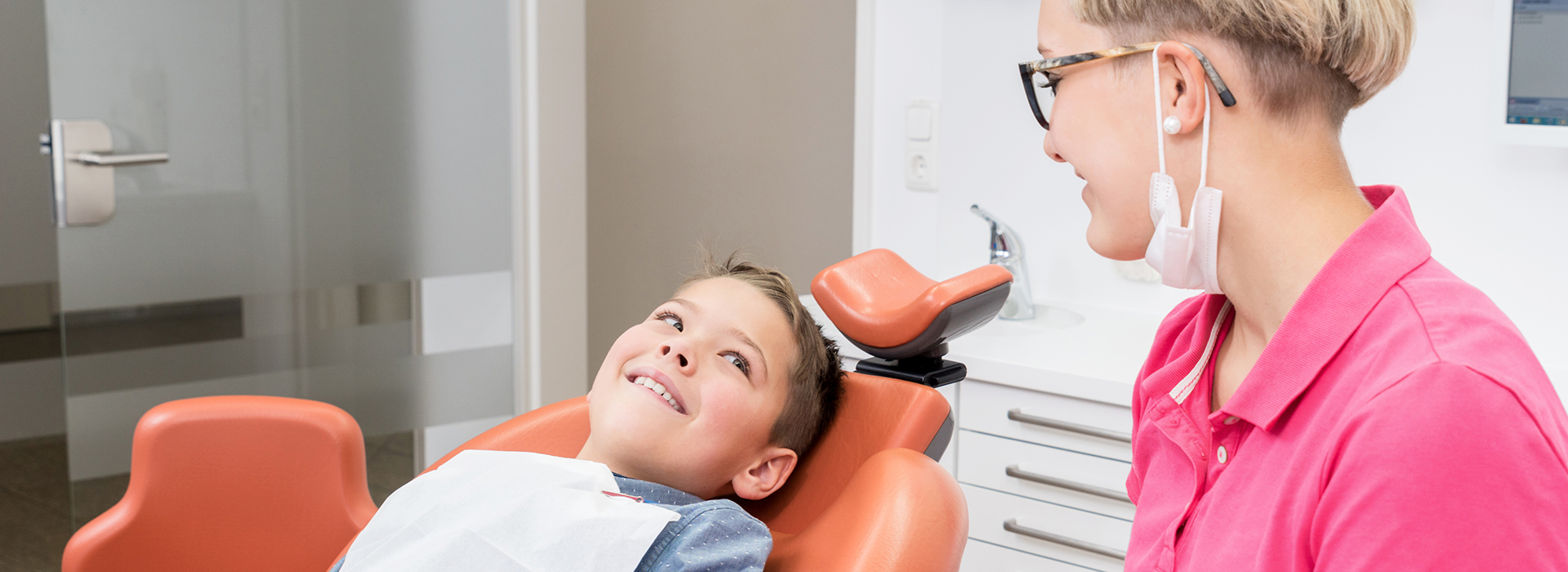 The image shows a dental setting where a young child is seated in a dental chair, receiving attention from a dental professional who appears to be conducting an examination or treatment.