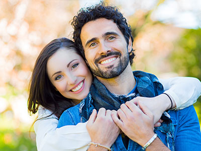 A man and a woman are hugging each other, smiling and appearing happy. The man is wearing a scarf around his neck and has a beard, while the woman has her hair tied back and is wearing a light-colored top. They are outdoors with greenery in the background.