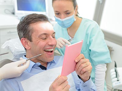 A man is sitting in a dental chair, holding a pink card or envelope, with a smiling woman in a surgical mask standing behind him.