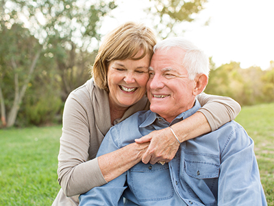 The image depicts an elderly couple hugging each other outdoors, with the man wearing a blue shirt and the woman in a white top.