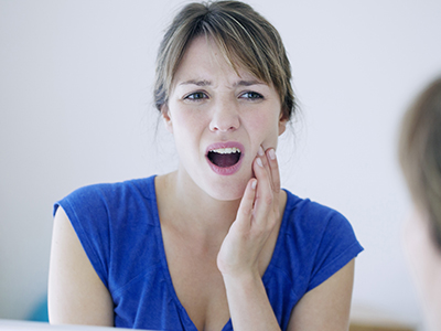 The image shows a woman with her mouth open, seemingly in the middle of speaking or singing, while looking at a mirror, which is reflecting her face. She appears to be indoors and has a concerned expression on her face.