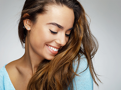 The image features a woman with long hair, smiling slightly and looking to the side. She has a fair complexion, wears light makeup, and is dressed in a blue top. The background is neutral and does not distract from the subject.