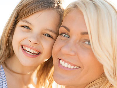 The image shows a smiling woman with blonde hair and a young girl with brown hair, both looking towards the camera. They appear to be outdoors during daylight.