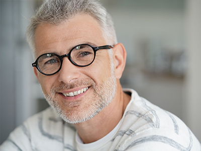 The image features a middle-aged man with short, graying hair, wearing glasses and a white shirt. He is smiling and looking directly at the camera.