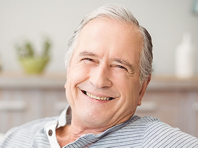 The image features an older man with white hair, wearing a blue shirt and smiling at the camera. He is seated comfortably in a chair indoors, looking relaxed and content.