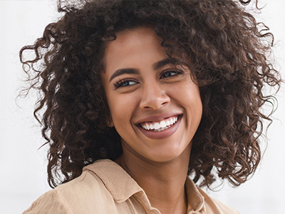The image shows a person with curly hair, smiling and looking slightly to the side. They have a joyful expression and are wearing a light-colored top.